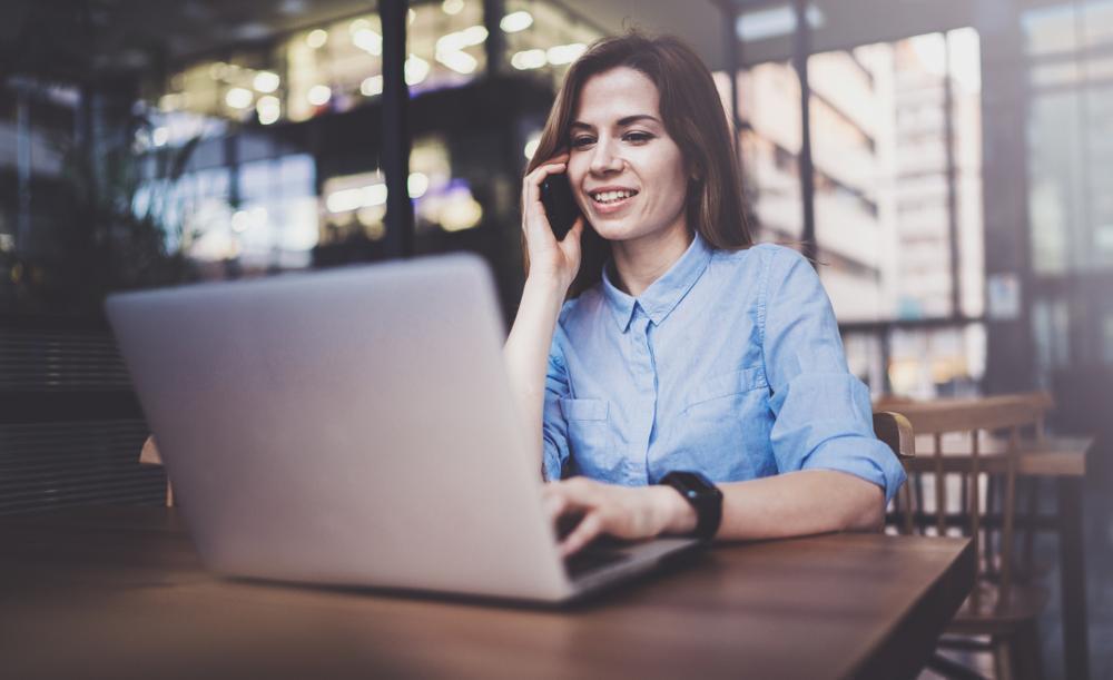 A woman working on her laptop while also talking on her phone.