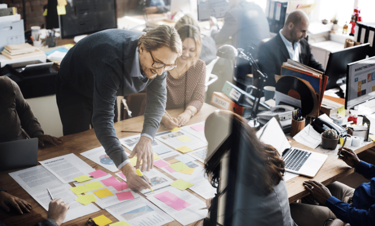 Employees smiling and working on documents sprawled out on a communal desktop. They are using highlighters and sticky notes.