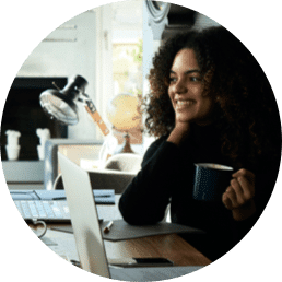 Circle photo of a woman holding a cup of coffee as she is listening to someone. She is sitting at a desk with her laptop open in front of her.