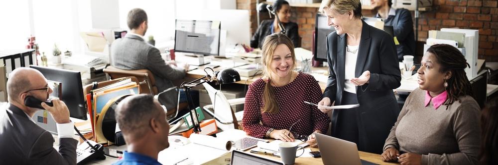 Office filled with employees talking and working. Woman standing with a piece of paper explaining to the rest of the employees. Everyone is looking happy.