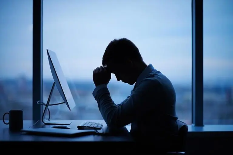 Man working on his computer in front of a large window. he has his head in his hands in sadness.