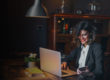 Woman in a darker office working on a laptop, smiling down at her cellphone