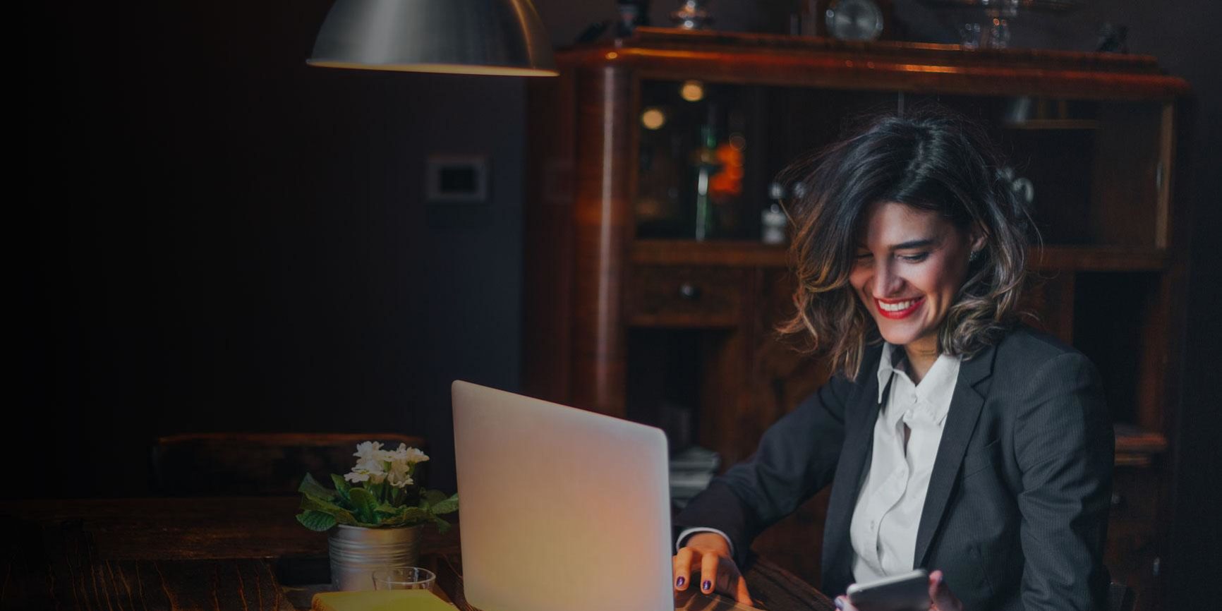 Woman in a darker office working on a laptop, smiling down at her cellphone