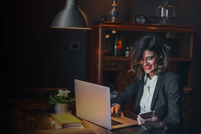 Woman in a darker office working on a laptop, smiling down at her cellphone