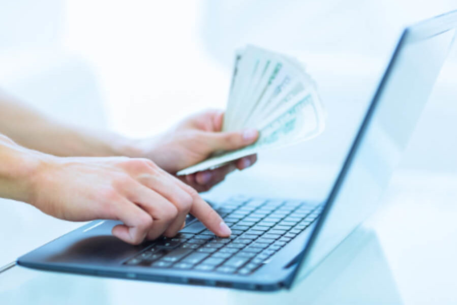 Close up of a person typing on a laptop with a handful of one hundred dollar bills in her other hand.