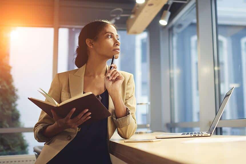 Woman holding a notebook at a desk with her laptop open. She is looking out the window in thought.