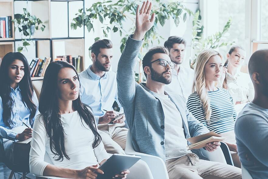 Employees sitting in a meeting listening and taking notes. A young gentlemen is raising his hand