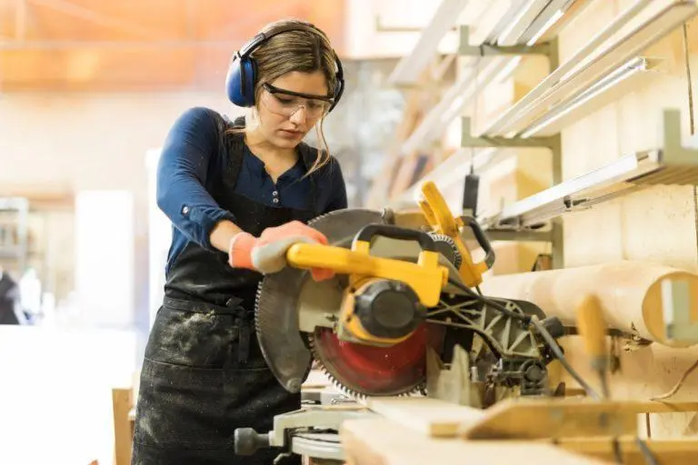 Woman with wood working protective gear on, sawing through a piece of wood.